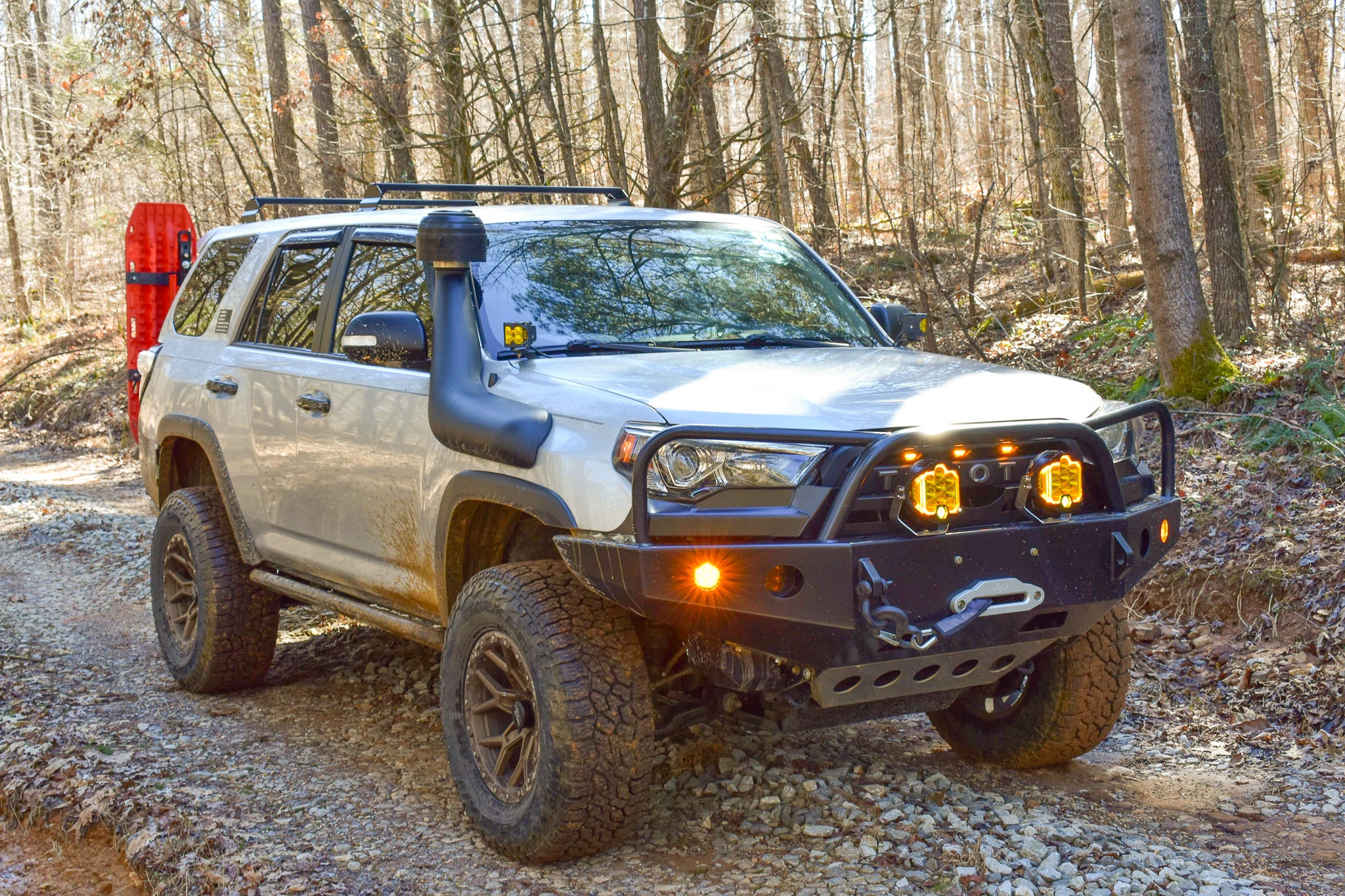 A silver 5th Gen 4Runner with a black minimalist overland rack mounted on top, driving through a wooded area.