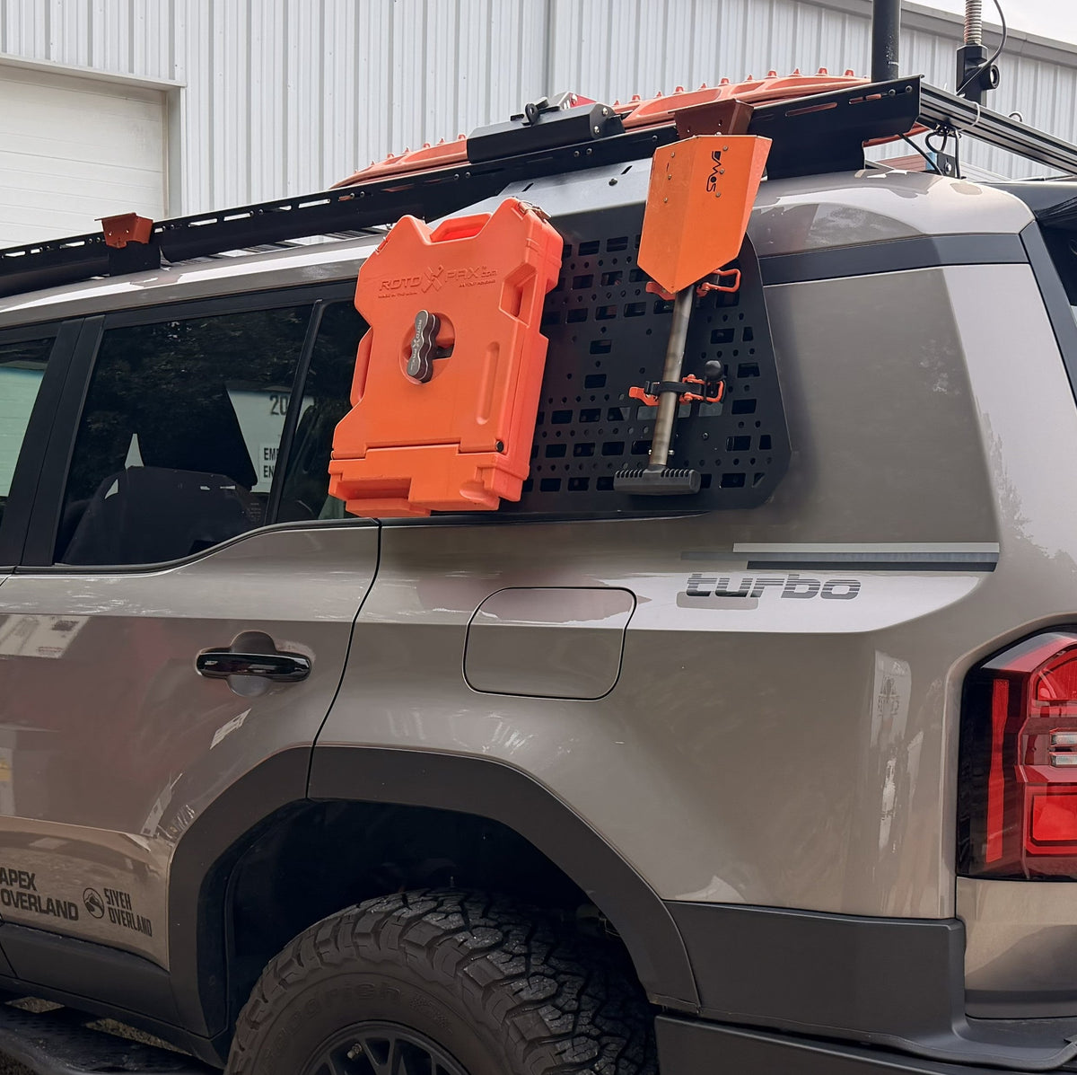 A gray Land Cruiser with off-road modifications, including a roof rack, orange storage bag, and rear utility rack, parked in front of a white industrial building.