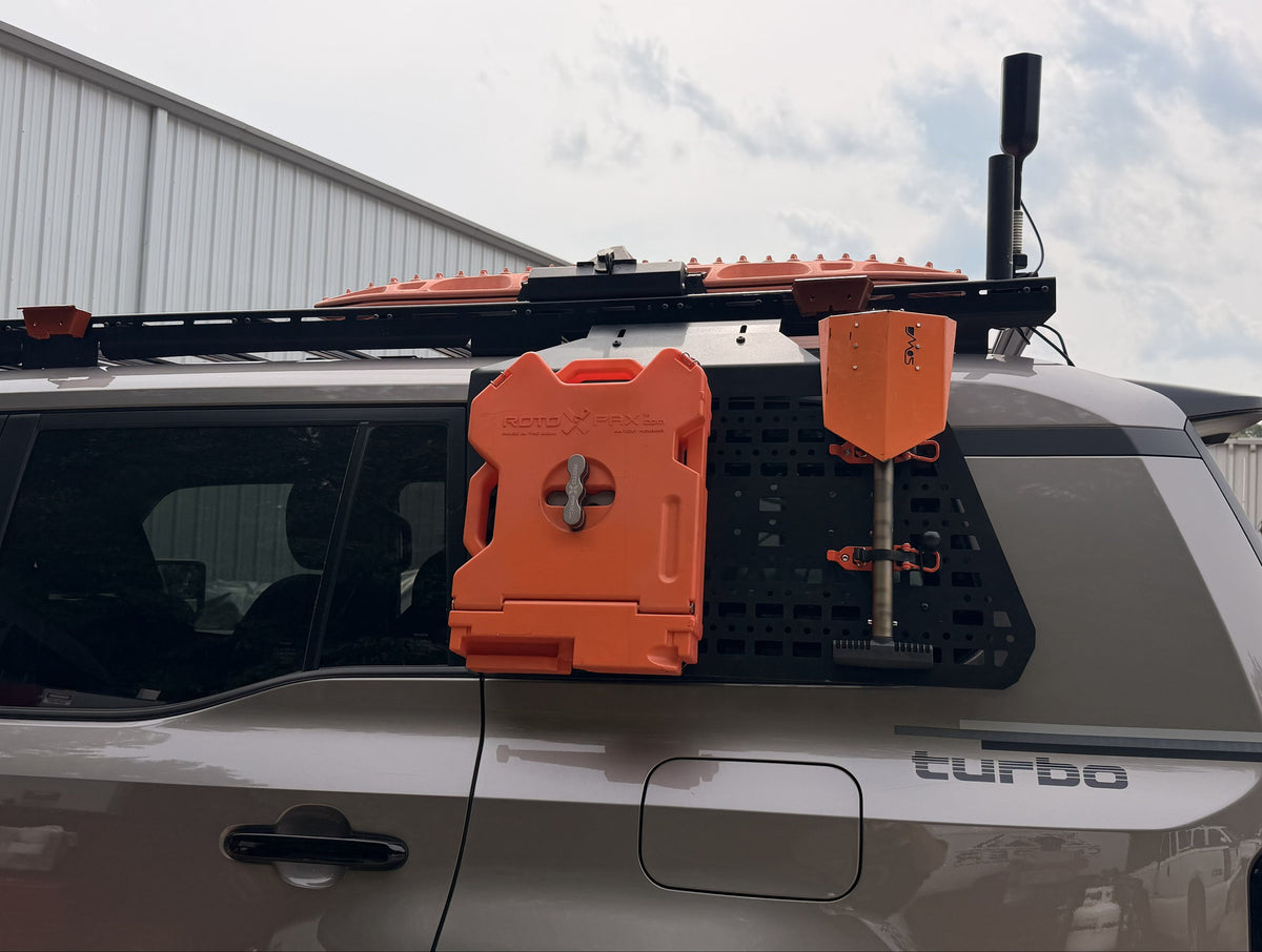 An orange rugged container and matching orange shovel mounted on the rear window rack of a gray SUV, photographed outdoors in front of a white building.