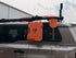 An orange rugged container and matching orange shovel mounted on the rear window rack of a gray SUV, photographed outdoors in front of a white building.