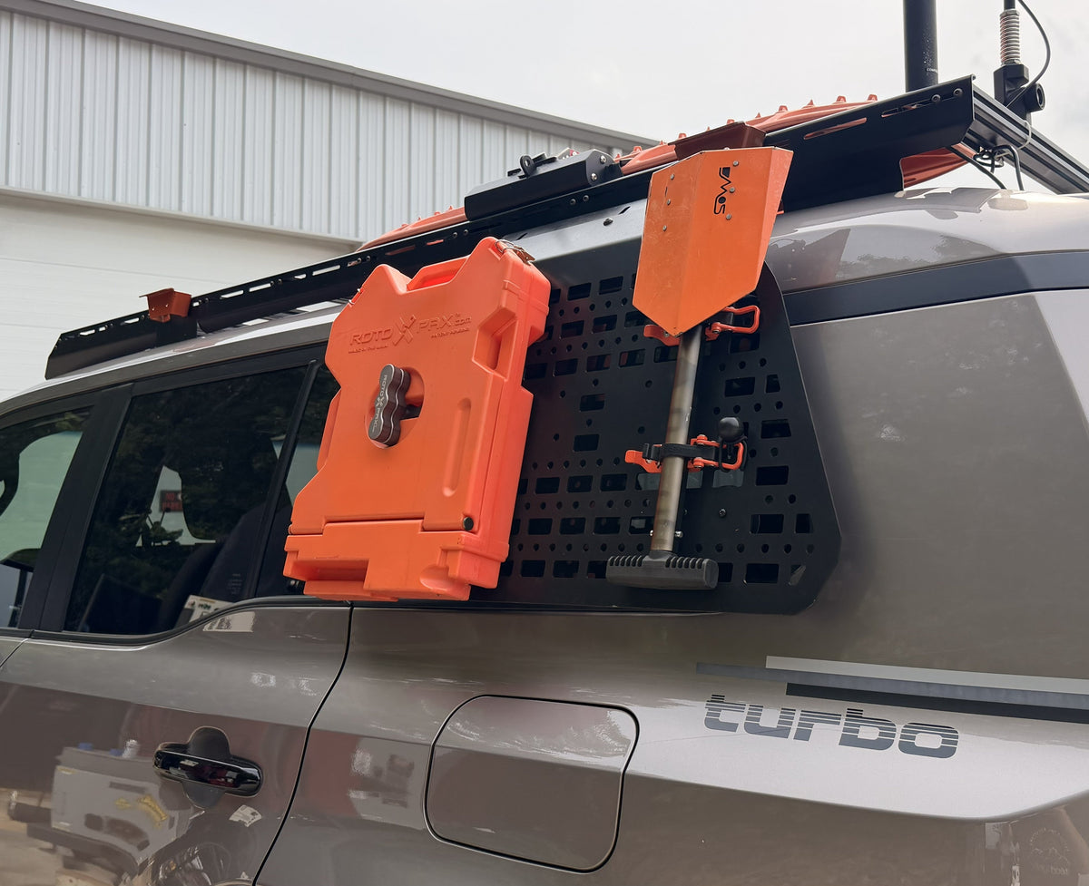 An orange emergency water container mounted on a black rack system on the rear side of a gray SUV, with an orange shovel also attached, photographed outdoors in front of a white garage building.