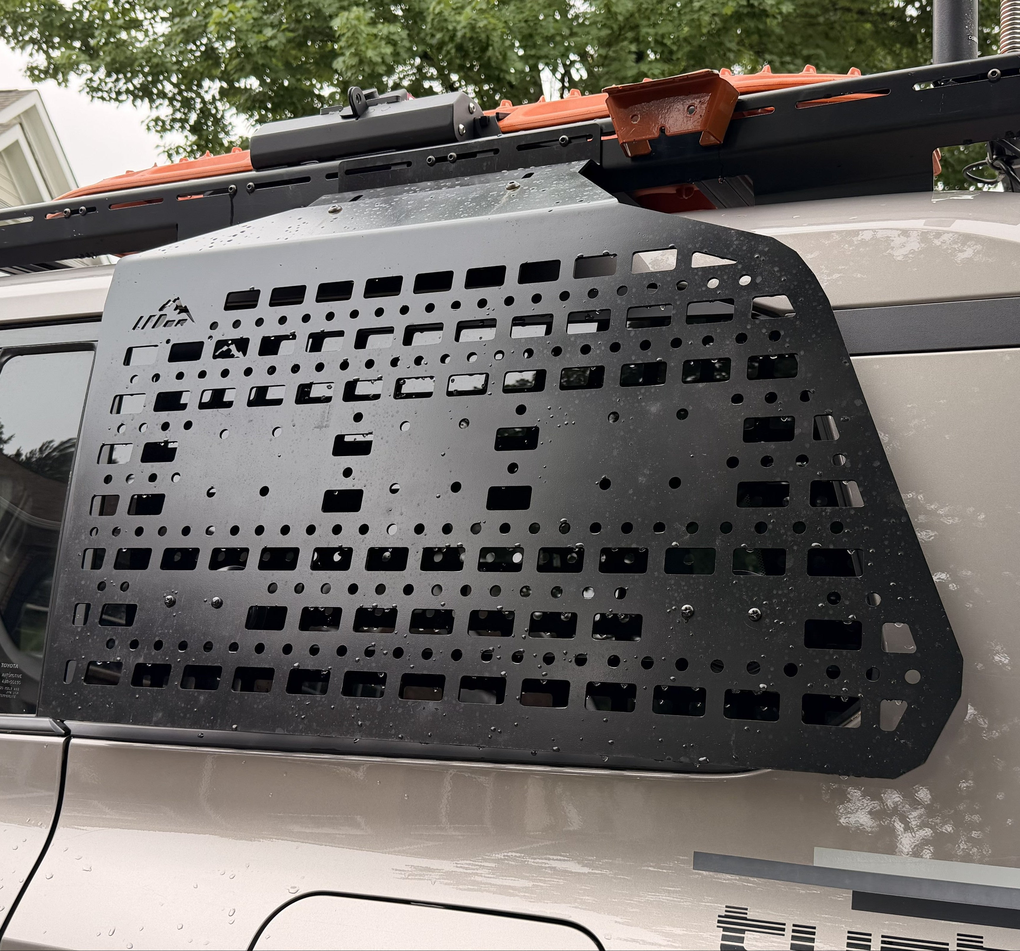 A black metal roof rack cargo carrier panel mounted on the rear window of a white vehicle, with trees and a house visible in the background.