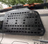 A black metal roof rack cargo carrier panel mounted on the rear window of a white vehicle, with trees and a house visible in the background.