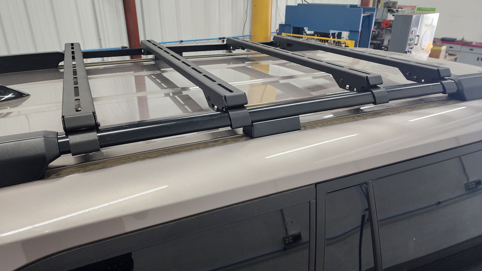 A black roof rack crossbar system mounted onto the roof of a light-colored vehicle, photographed in an indoor workshop or garage setting.