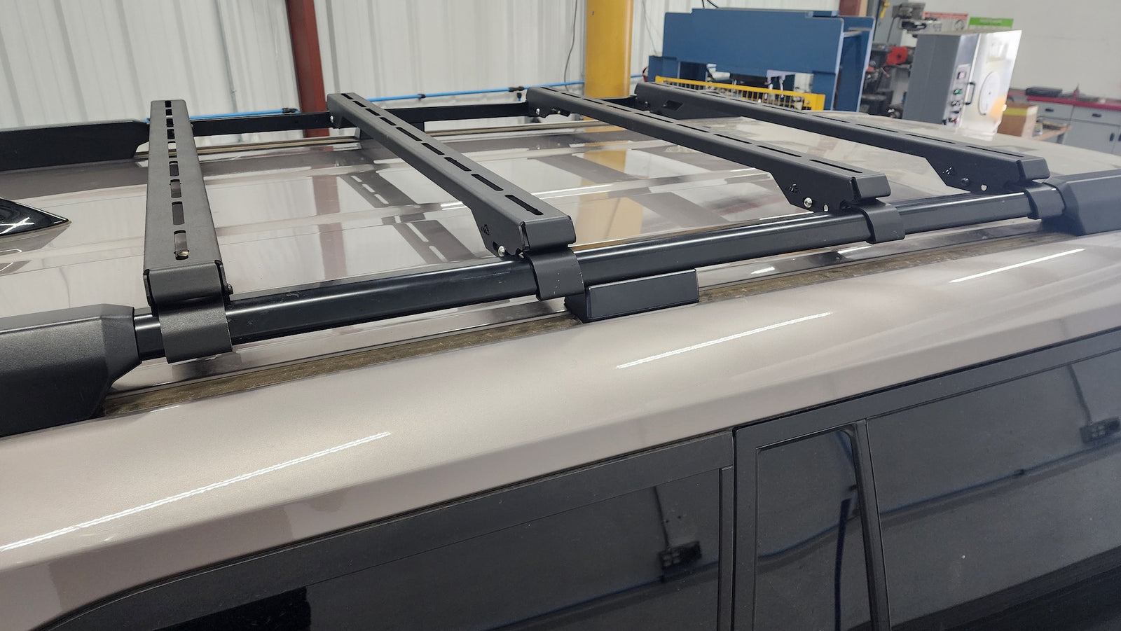 A black roof rack crossbar system mounted onto the roof of a light-colored vehicle, photographed in an indoor workshop or garage setting.