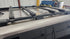 A black roof rack crossbar system mounted onto the roof of a light-colored vehicle, photographed in an indoor workshop or garage setting.