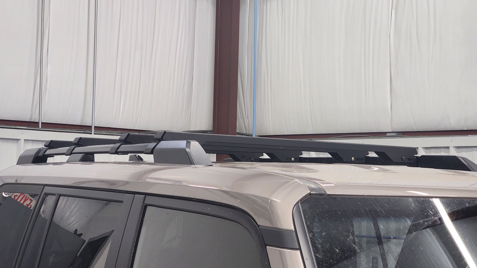 A black roof rack crossbar system mounted on the roof of a beige SUV, photographed in a warehouse or garage setting with a corrugated metal wall in the background.