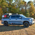 A silver Toyota Tundra CrewMax truck with a roof rack installed, parked in a natural setting with trees in the background.