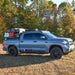 A silver Toyota Tundra CrewMax truck with a roof rack installed, parked in a natural setting with trees in the background.