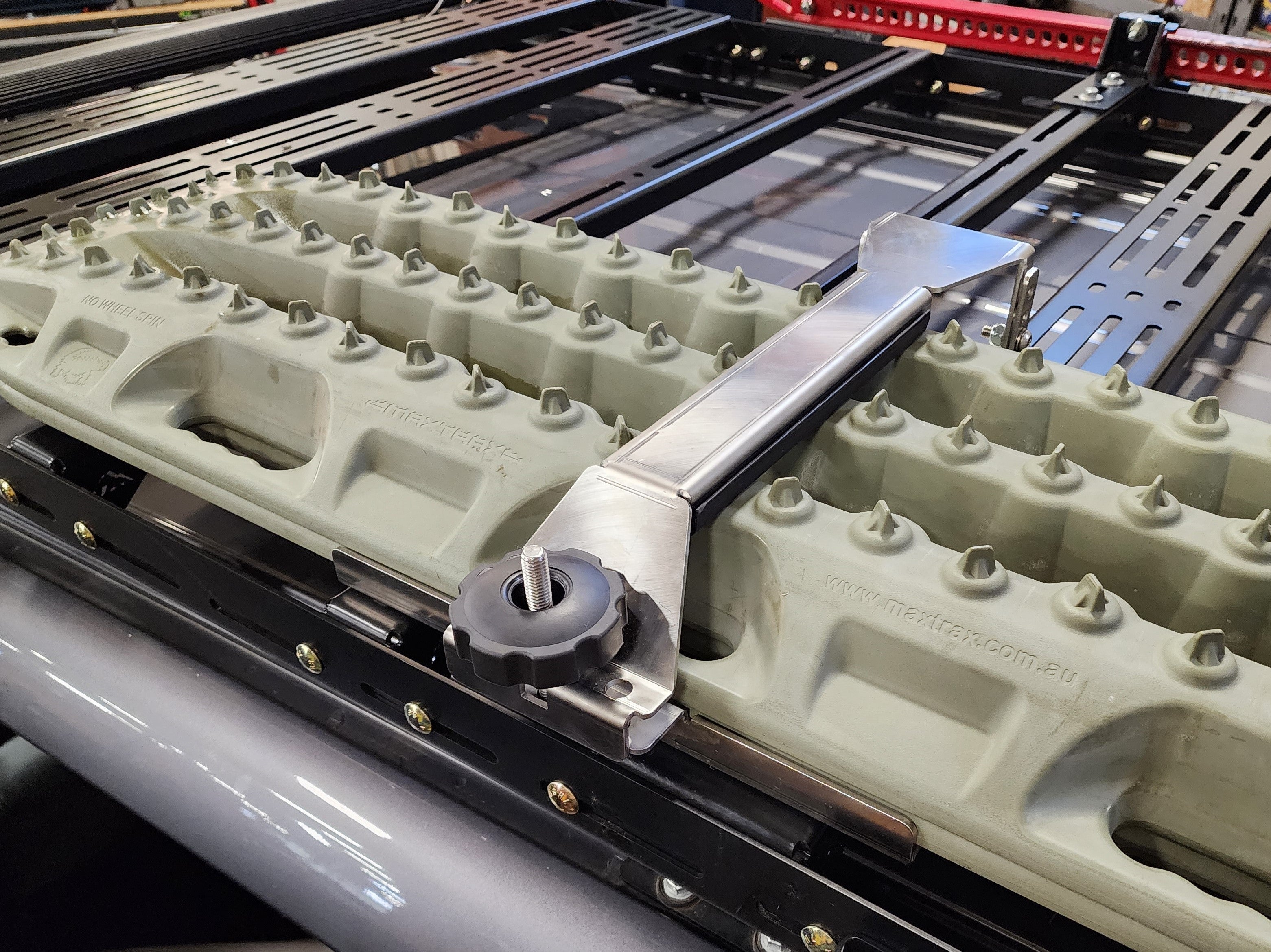 A stainless steel traction board mount bolted to a roof rack, with a raw finish.