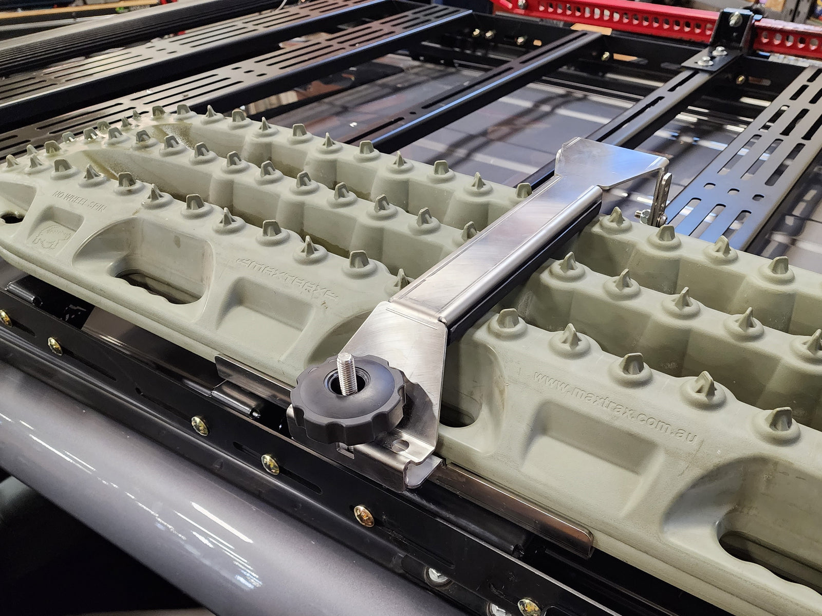 A stainless steel traction board mount bolted to a roof rack, with a raw finish.