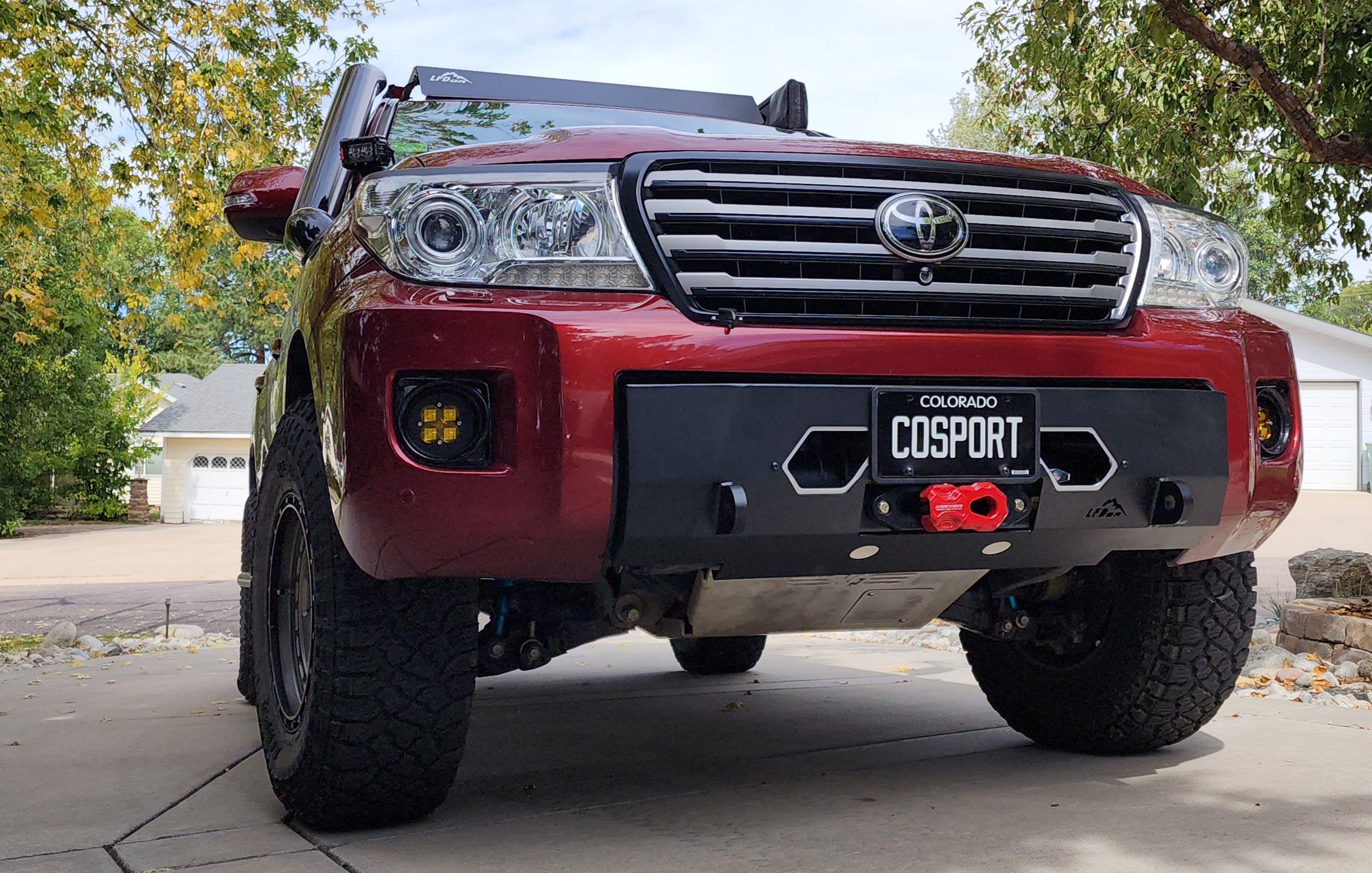 Front bumper for Land Cruiser 200 featuring a winch mount, recovery points, and lower grille, displayed on a red vehicle.
