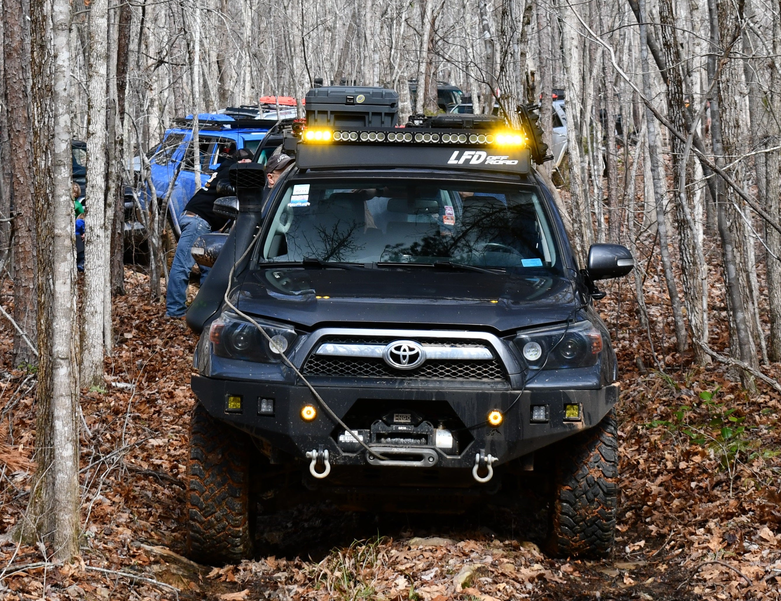 A black 5th Gen 4Runner with a curved front crossbar for full racks, seen driving through a wooded area.