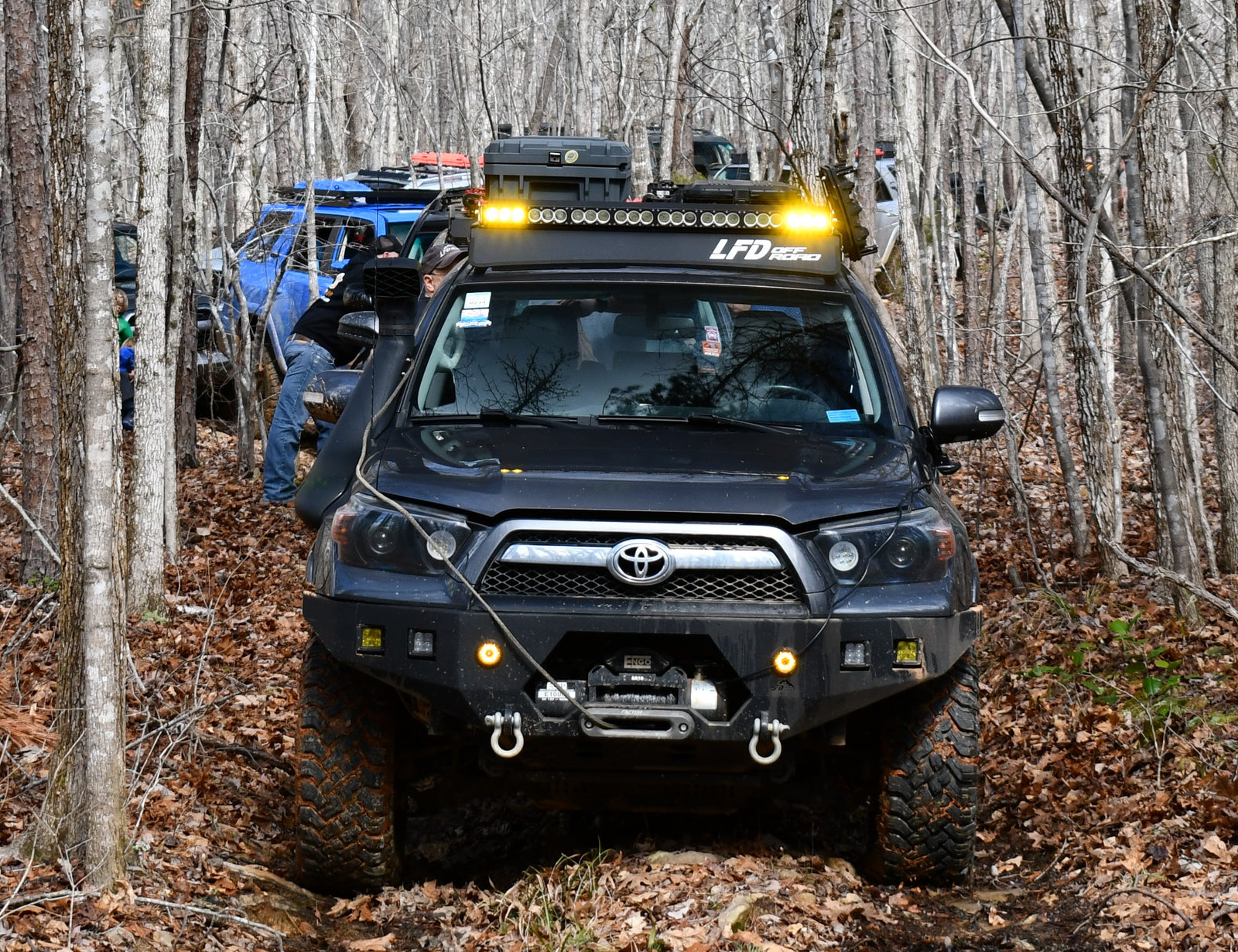 A black 5th Gen 4Runner with a curved front crossbar for full racks, seen driving through a wooded area.