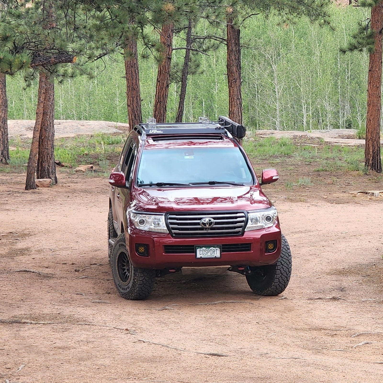 A 200 Series Land Cruiser SUV with a set of crossbars installed on the roof rails, located in a natural outdoor setting.