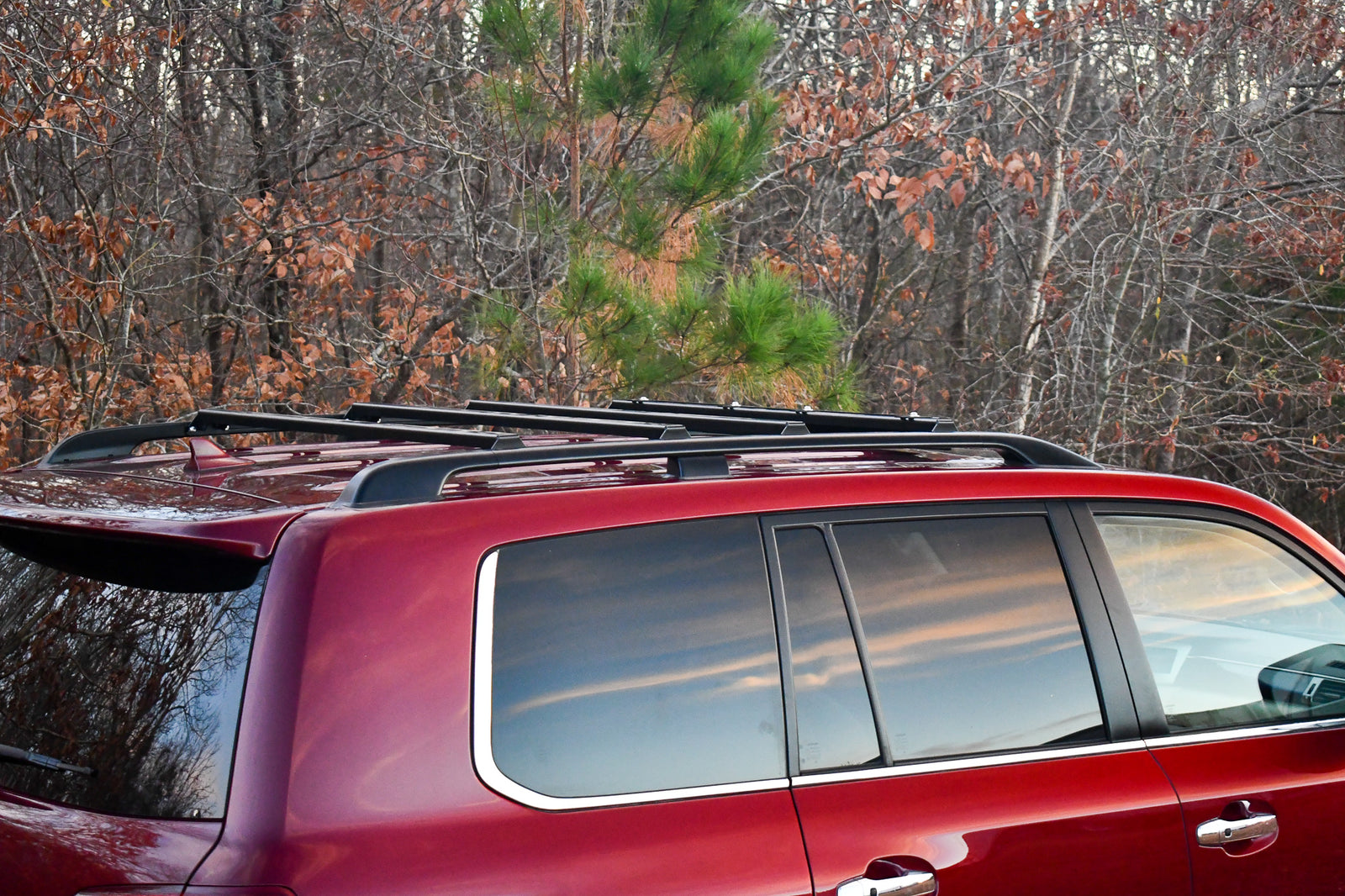 Ruggedized Crossbars mounted on the roof rails of a red 200 Series Land Cruiser vehicle, set against a forest backdrop.