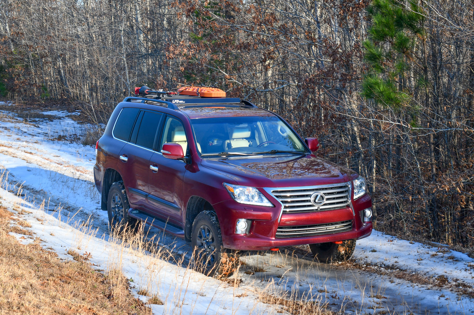 A red Lexus LX570 SUV with factory side rails and a set of black ruggedized crossbars installed on top, parked in a wooded area with snow on the ground.