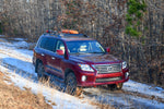 A red Lexus LX570 SUV with factory side rails and a set of black ruggedized crossbars installed on top, parked in a wooded area with snow on the ground.