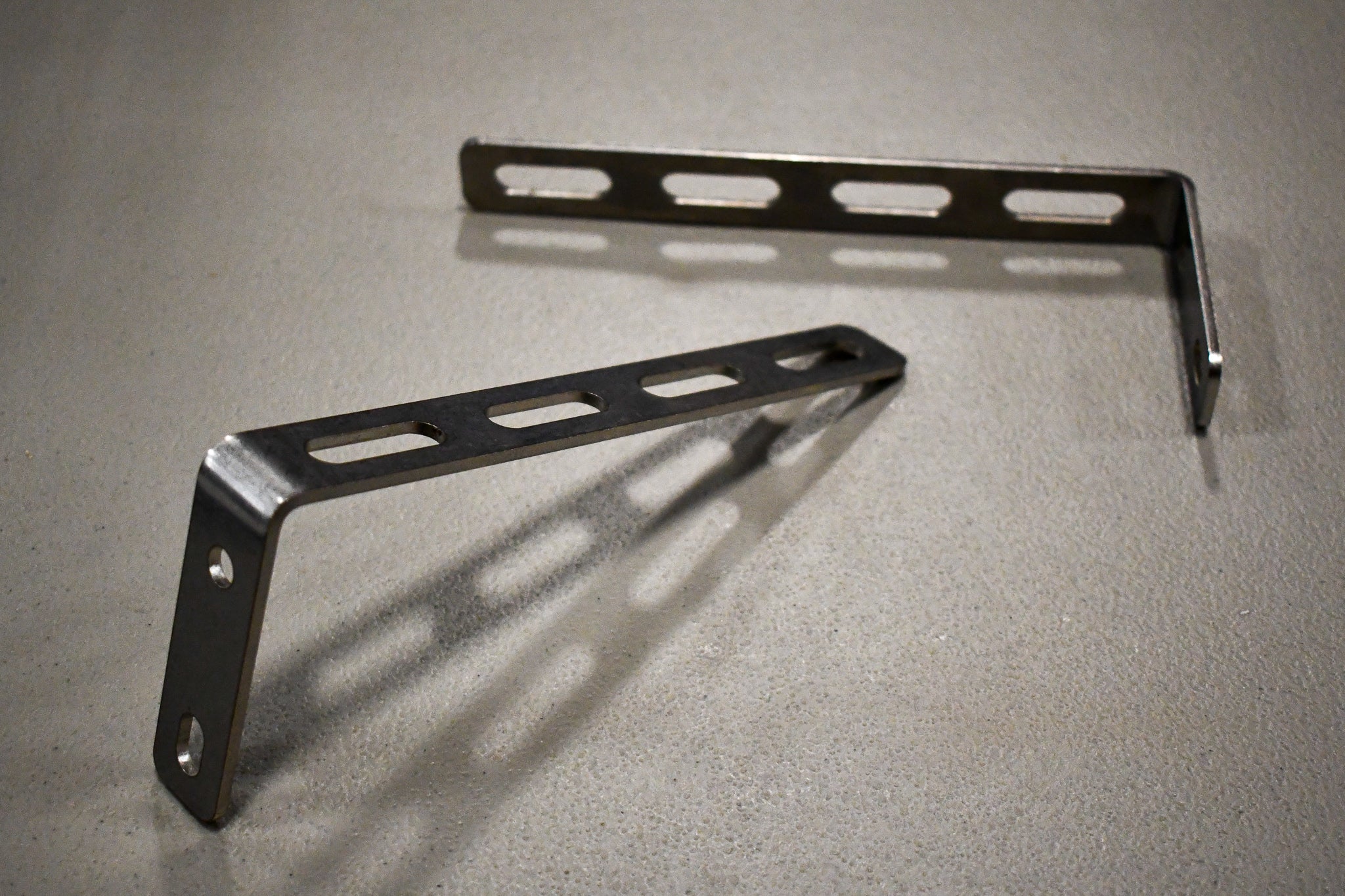 A pair of stainless steel universal awning mounts with multiple bolt holes, designed to be mounted on a vehicle roof rack.