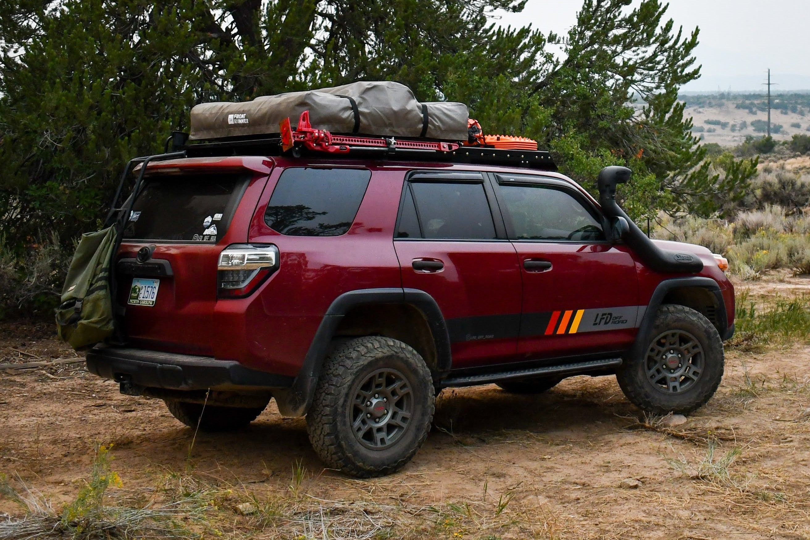 A red SUV with a roof rack that includes an angled Hi-Lift mount, allowing a Hi-Lift jack to be mounted on the side of the rack.
