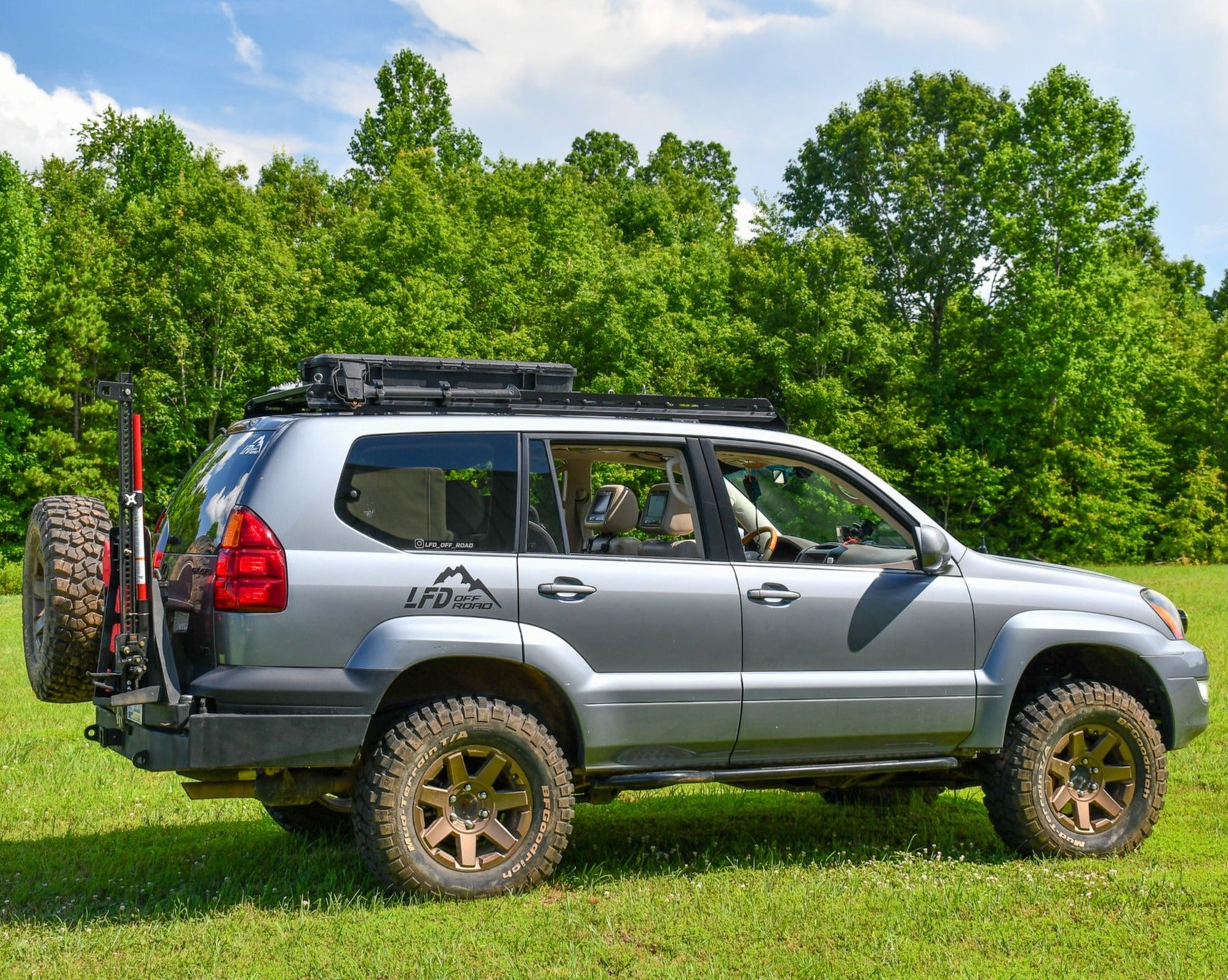 A GX470 Lexus SUV equipped with a full roof rack featuring ruggedized crossbars and an integrated wind fairing crossbar, displayed in a natural setting with trees in the background.