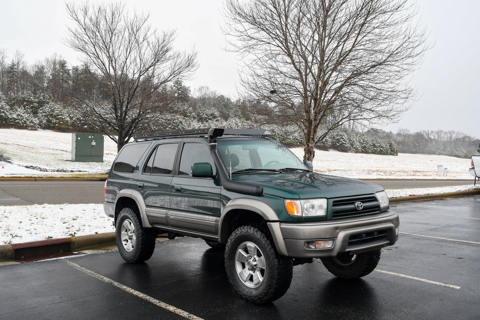 A green 3rd Gen 4Runner with a full steel roof rack installed on top, parked in a lot with trees and a building in the background.