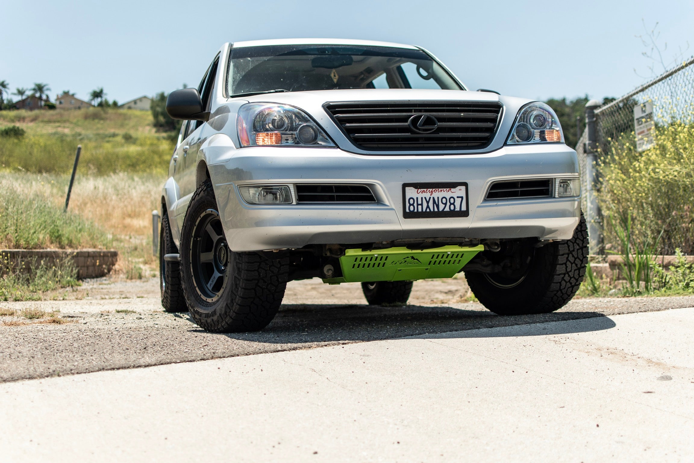 A silver Lexus GX470 with a green front skid plate installed on the vehicle, positioned on a road with a natural landscape in the background.