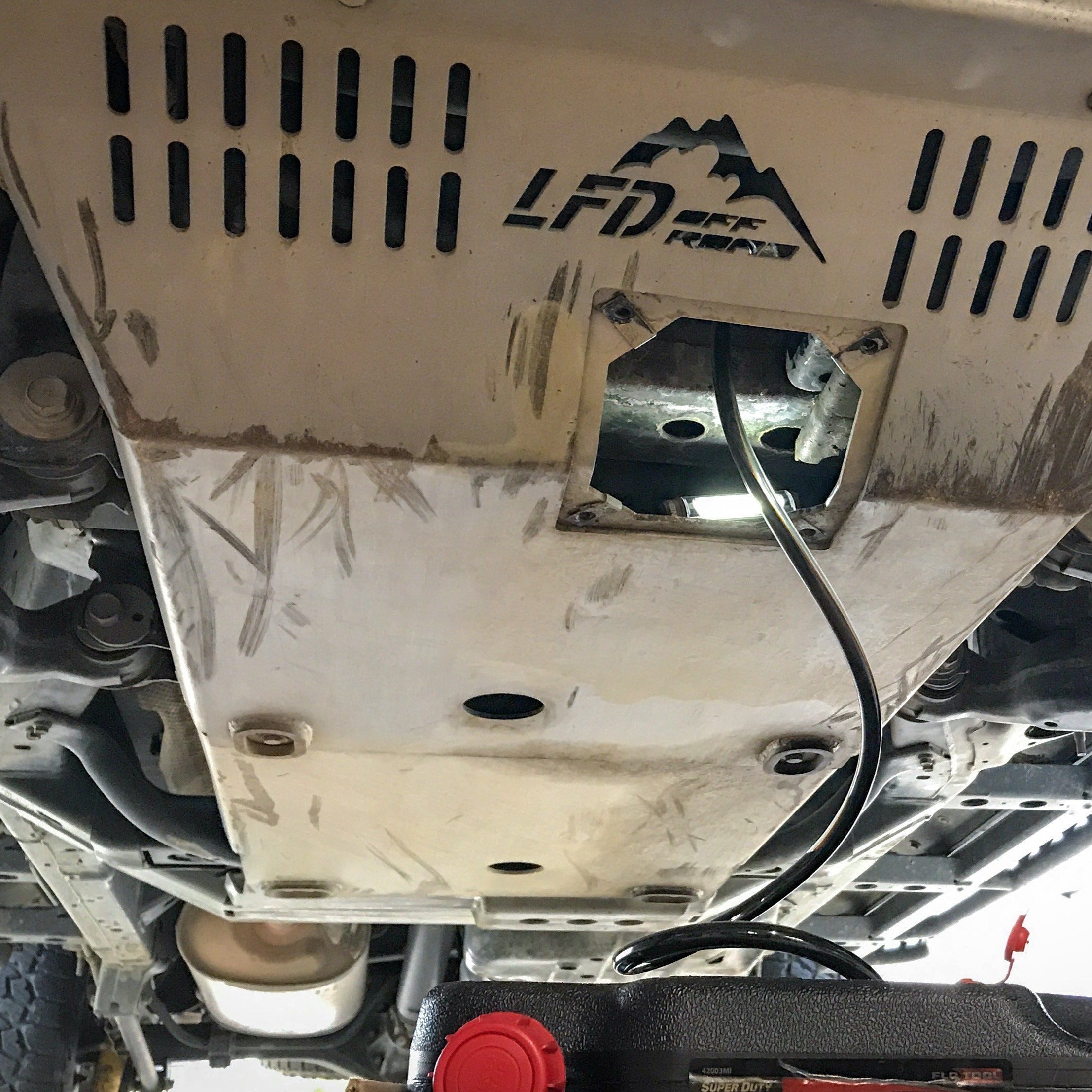 A photo showing the underside of a vehicle with a focus on a metal skid plate designed for off-road vehicle modifications. The image displays the placement of the skid under the vehicle's engine area.