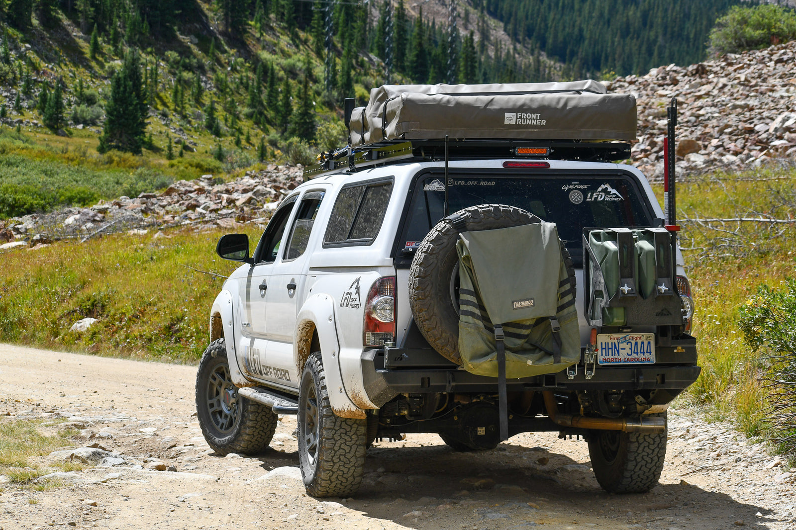 A white 2nd Gen Tacoma with a no cut rear bumper and off-road tires, parked in a scenic outdoor setting with trees in the background.