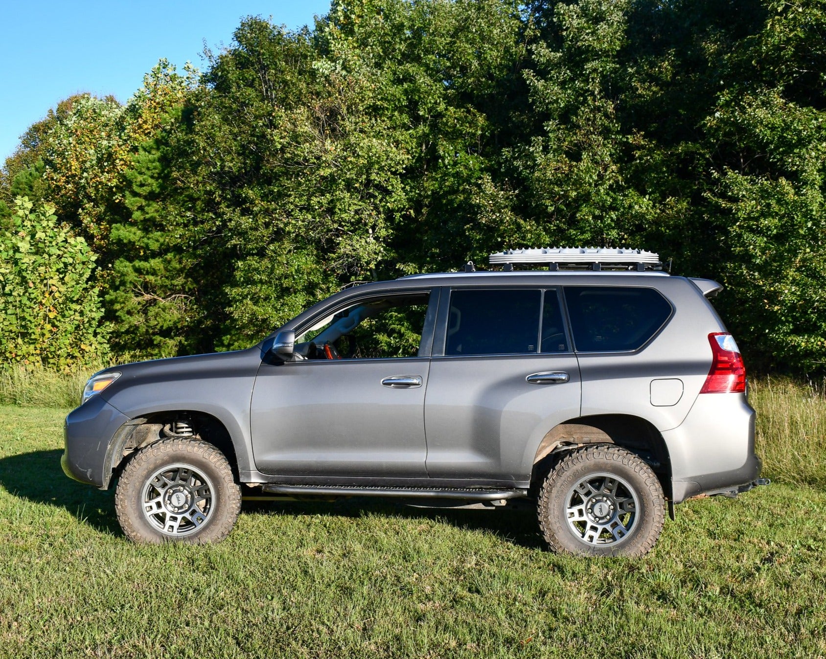 A GX460 vehicle with rock sliders installed, positioned in a natural outdoor setting with trees in the background.