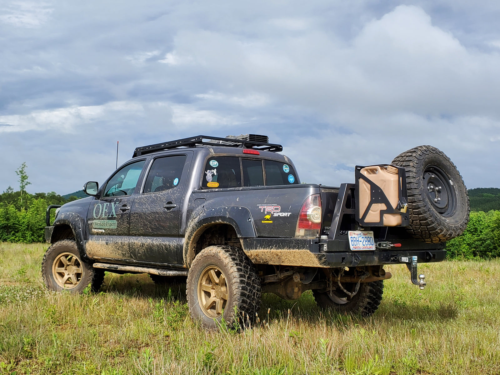 A black single jerry can mount attached to the rear bumper of a vehicle, set against a outdoor backdrop.