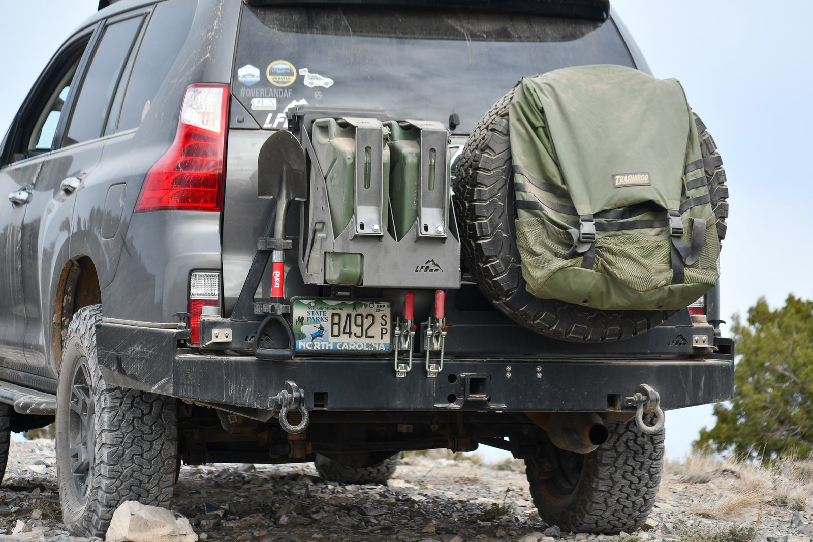 A dual jerry can mount attached to the rear bumper of a vehicle, with one canister partially visible and the other removed, revealing the mounting mechanism.